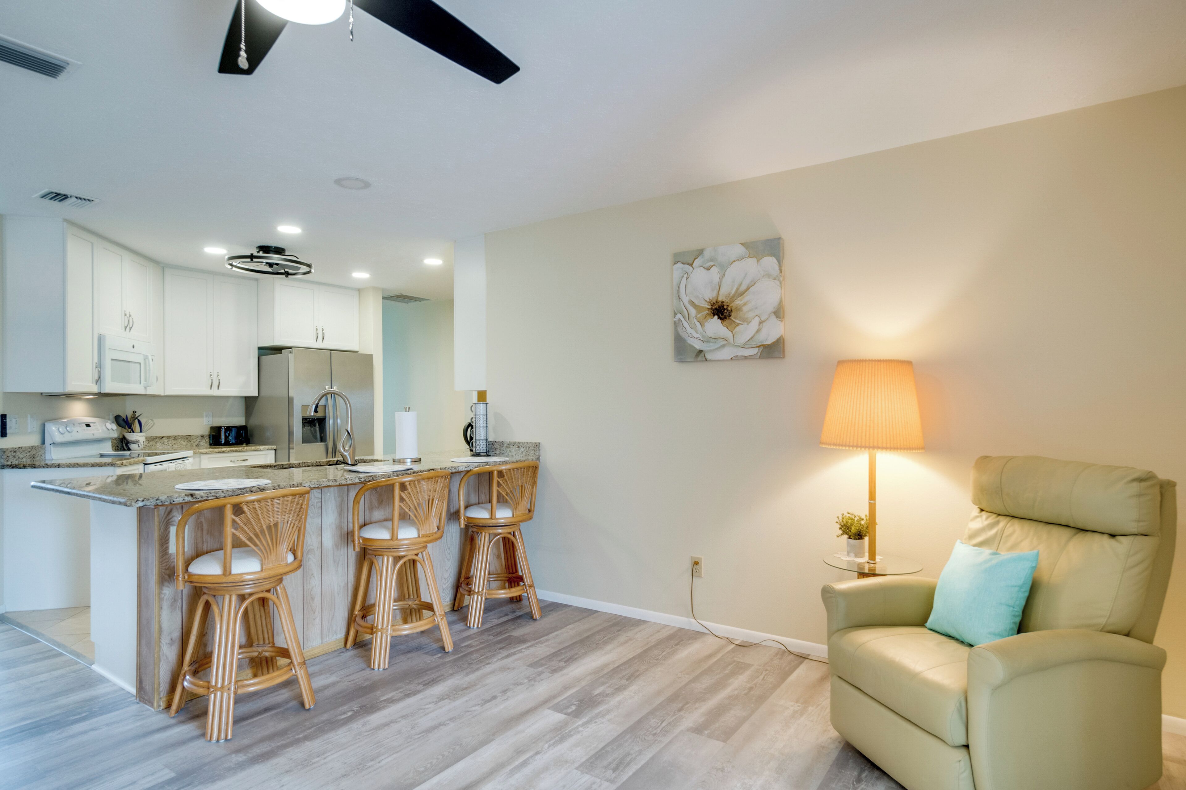 A modern kitchen with white cabinets, a central island with bar stools, a dining area with a table and chairs, and a living room with a sofa and a lamp.