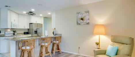 A modern kitchen with white cabinets, a central island with bar stools, a dining area with a table and chairs, and a living room with a sofa and a lamp.