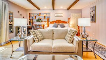 A cozy hotel room with a sofa, a glass coffee table, a bed with pillows, a nightstand with a lamp, a bookshelf, and a painting on the wall.