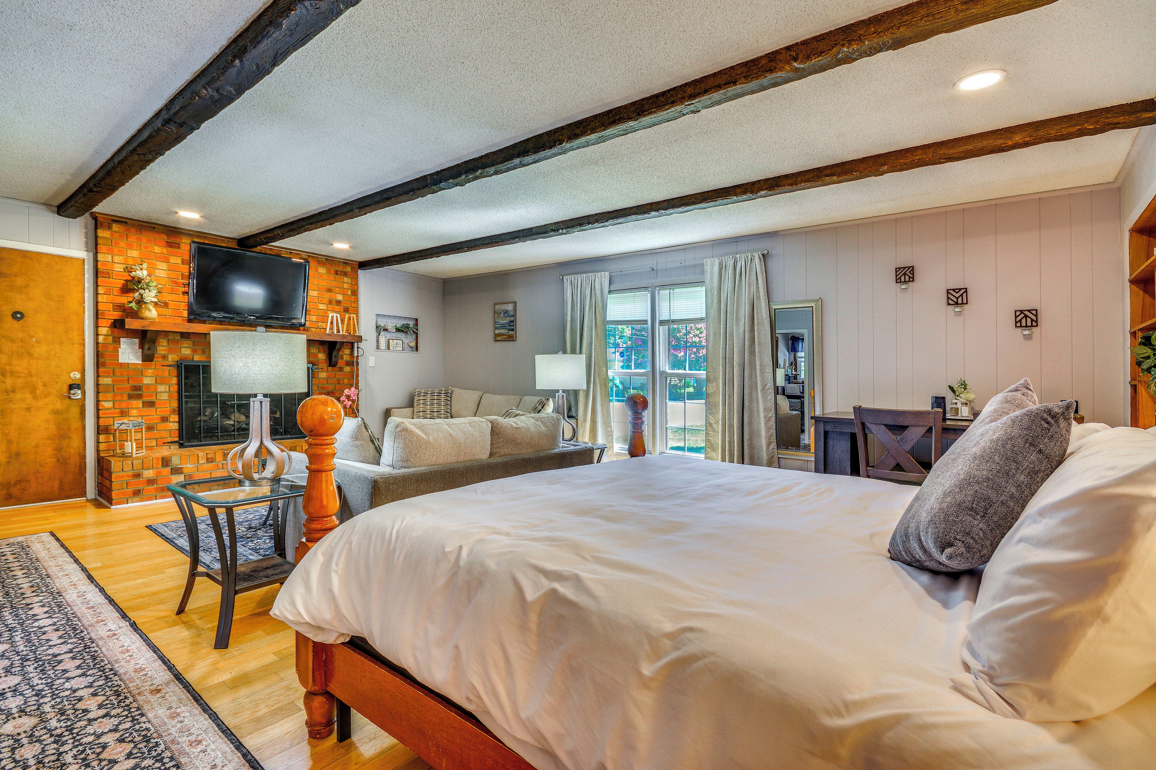 A hotel room with a large bed, a television mounted on a brick wall, a fireplace, and a view of the outdoors through a window.