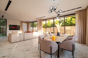 A modern living room with a sofa set, a dining table with chairs, a TV mounted on the wall, and large windows. - Villa Vamo | Luxury 5-Bedroom Golf Villa with Heated Pool | Amelkis Marrakech (Marrakech)