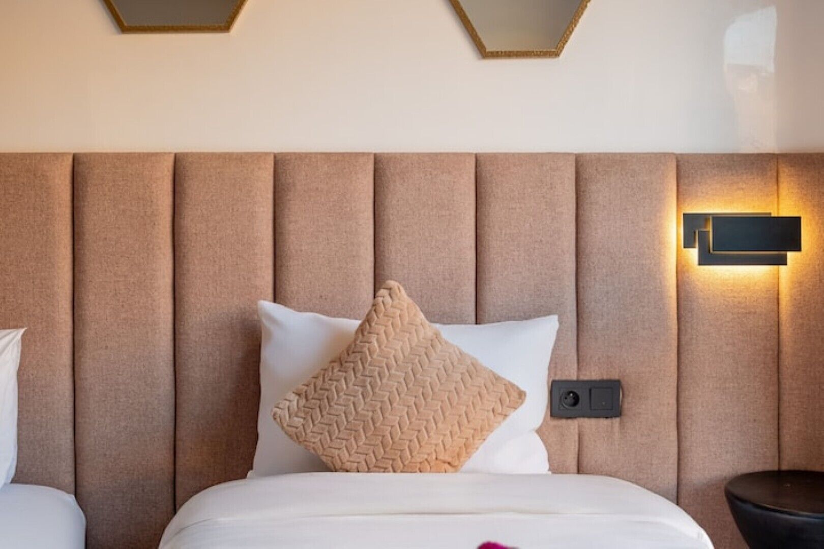 A hotel room with a padded headboard, a bed with white bedding, a beige pillow, and a wall-mounted light fixture.