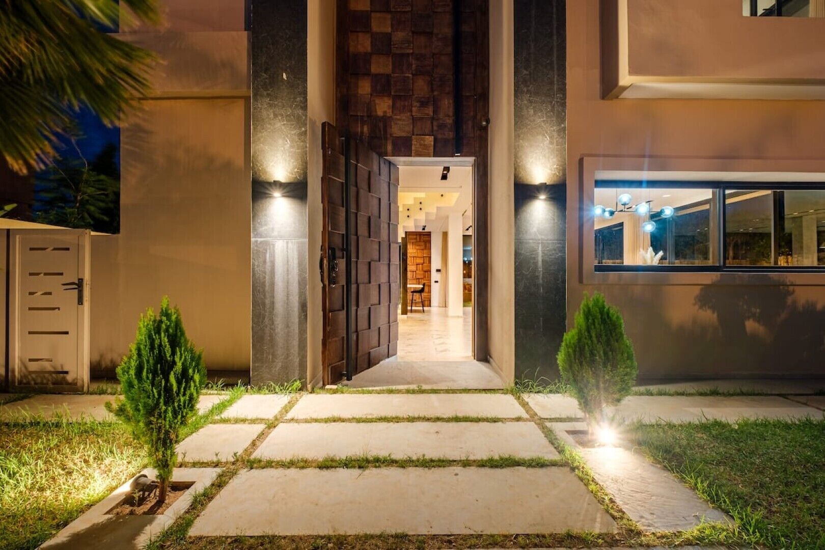 A modern house entrance with a pathway leading to a door, a small tree, and a window with lights.