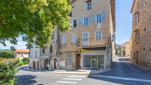A street view with a building featuring a ground-floor shop with a sign, a balcony, and a crosswalk in front. - La Vague – Comfortable Apartment in the Heart of Montauroux Village (Montauroux)