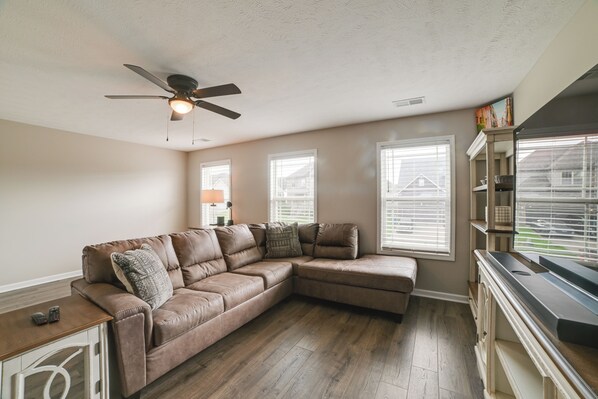 A living room with a sectional sofa, a ceiling fan, and a view of the outside through the windows.