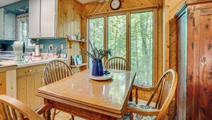 A wooden interior with a dining table, chairs, a clock, and a window with a view of trees.
