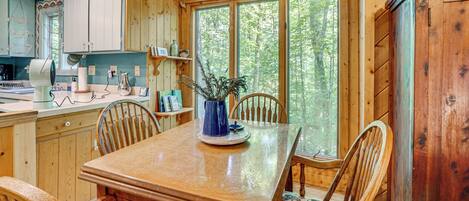 A wooden interior with a dining table, chairs, a clock, and a window with a view of trees.