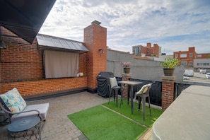 Eine Dachterrasse mit Backsteinmauer, einer überdachten Sitzgelegenheit, einem kleinen Tisch und einem grünen Kunstturf.