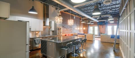 Una cocina moderna con electrodomésticos de acero inoxidable, una isla central con taburetes de bar, iluminación colgante y una pared decorativa de ladrillo.