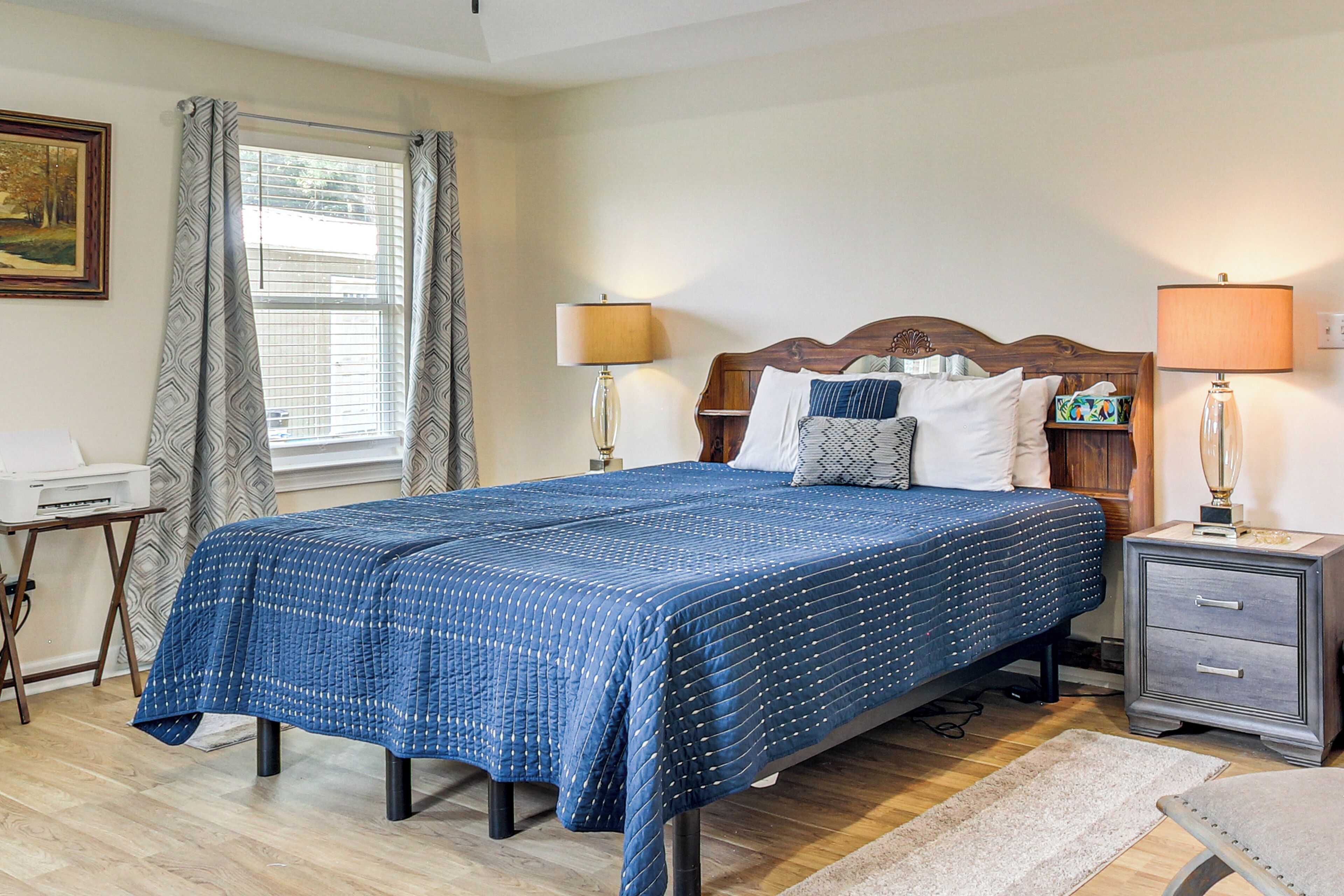 A neatly made bed with a blue quilt, two bedside lamps, a nightstand with a lamp, a wooden headboard, a framed picture on the wall, and a window with curtains.
