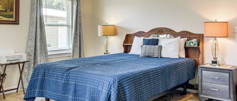 A neatly made bed with a blue quilt, two bedside lamps, a nightstand with a lamp, a wooden headboard, a framed picture on the wall, and a window with curtains.