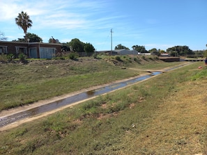 Un paysage rural avec un canal d’eau, des zones herbeuses et quelques bâtiments en arrière-plan.