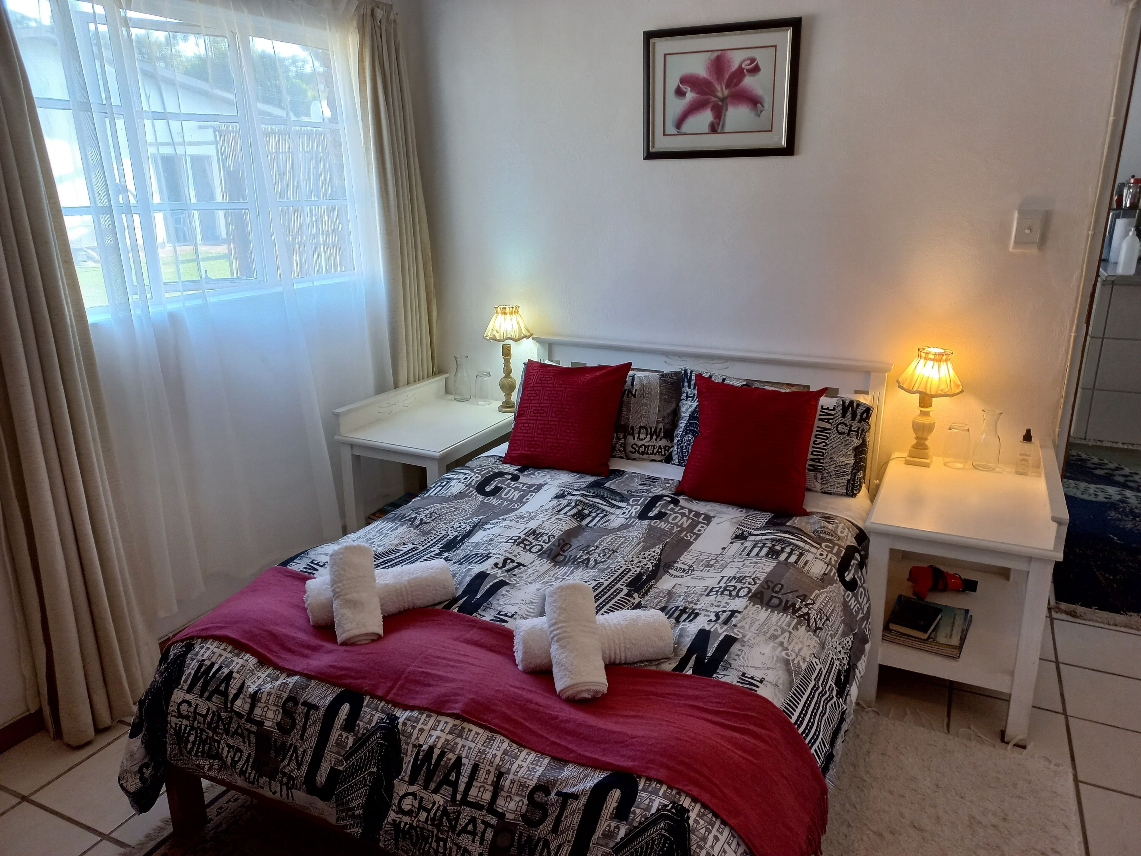 A neatly made bed with a patterned bedspread, two red pillows, and two white towels folded on top. There are two bedside tables with lamps, a framed picture on the wall, and a window with curtains.