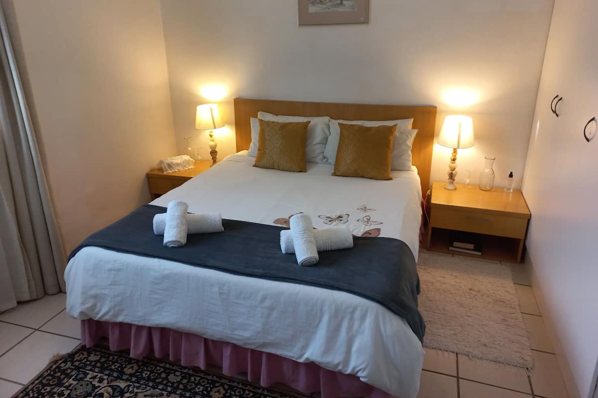 Una cama bien tendida con una colcha blanca y azul oscuro, dos almohadones decorativos y dos toallas enrolladas sobre la cama. Hay dos lámparas de noche en mesitas de madera, un cuadro enmarcado en la pared sobre la cama y una alfombra estampada en el suelo.