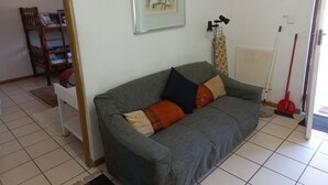 Una sala de estar con un sofá gris, una obra de arte enmarcada, una silla de madera y una mopa apoyada en la pared.