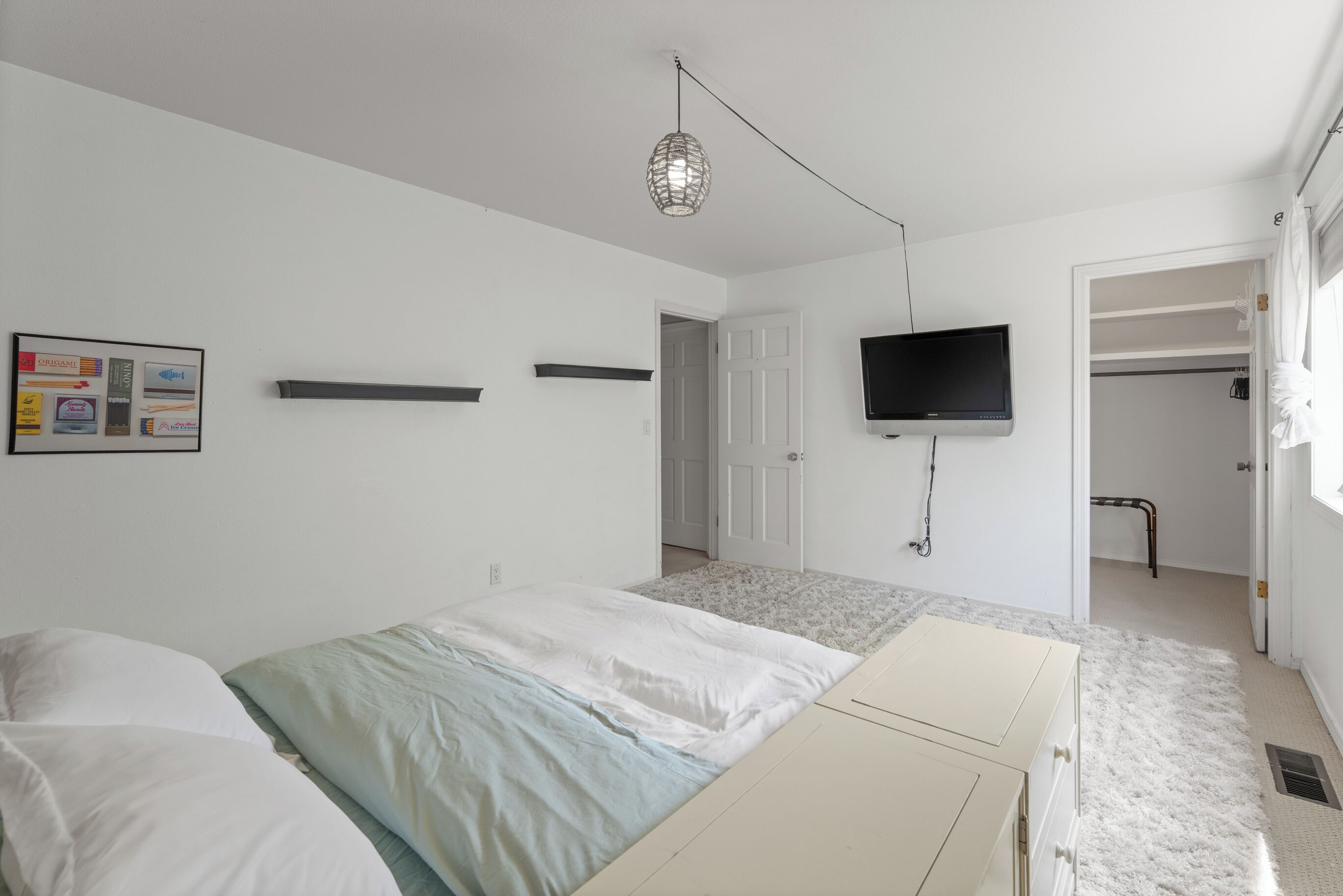 Un dormitorio con una cama, un televisor montado en la pared, una puerta que da a un baño y un mueble pequeño.