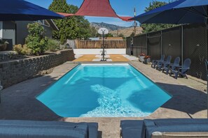 Ein rechteckiger Pool mit Sprungbrett, umgeben von Liegestühlen und Sonnenschirmen, vor einer Hügellandschaft und einem Holzzaun.