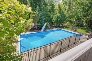 Uma piscina retangular com um escorrega, cercada por uma grade de metal e árvores ao fundo.