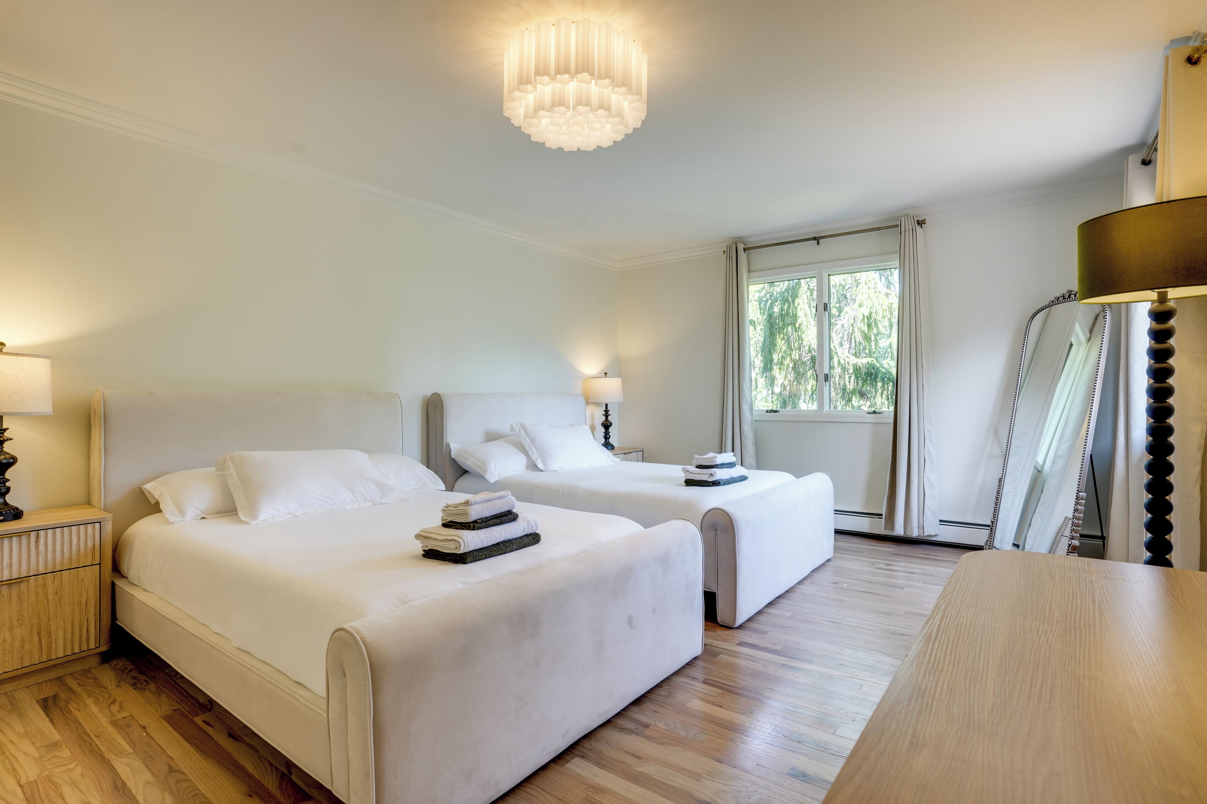 A hotel room with two beds, a wooden floor, a chandelier, and a window with curtains.