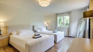 A hotel room with two beds, a wooden floor, a chandelier, and a window with curtains.