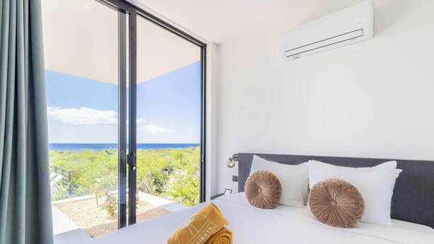 Une chambre d’hôtel avec un lit, deux oreillers, une serviette roulée, un appareil de climatisation et une vue sur la verdure et la mer.