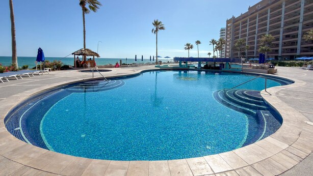 A swimming pool with a curved design, surrounded by palm trees and a beach view. - Wake Up to the Sea Oceanfront Retreat W207 (Puerto Peñasco)