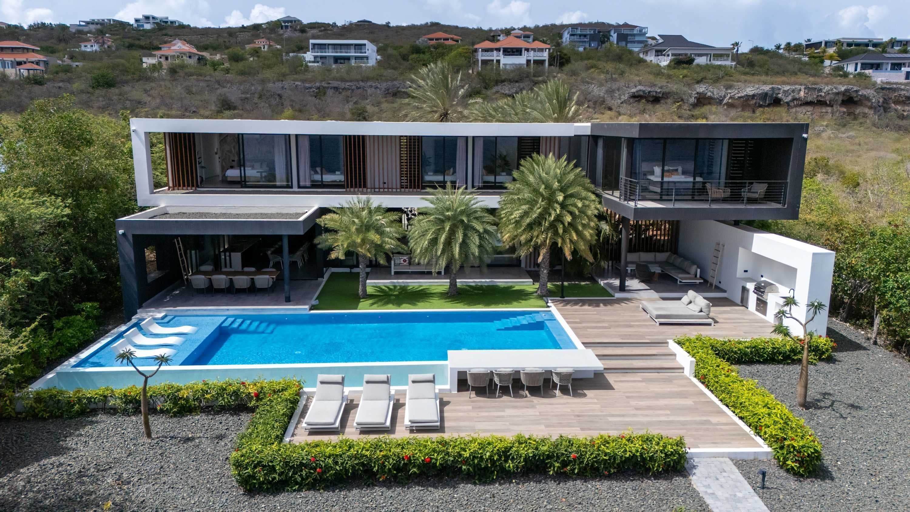 Ein modernes, zweistöckiges Haus mit Pool, Sitzgelegenheiten im Freien und Blick auf die umliegende Landschaft.