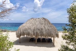 Eine Strandhütte mit einem Reetdach, umgeben von Sand und freiem Blick auf den Ozean.