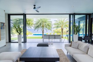 Ein modernes Wohnzimmer mit einem großen Glasfenster, das zu einem Pool und Meerblick führt, einem Deckenventilator und einer minimalistischen Möblierung.