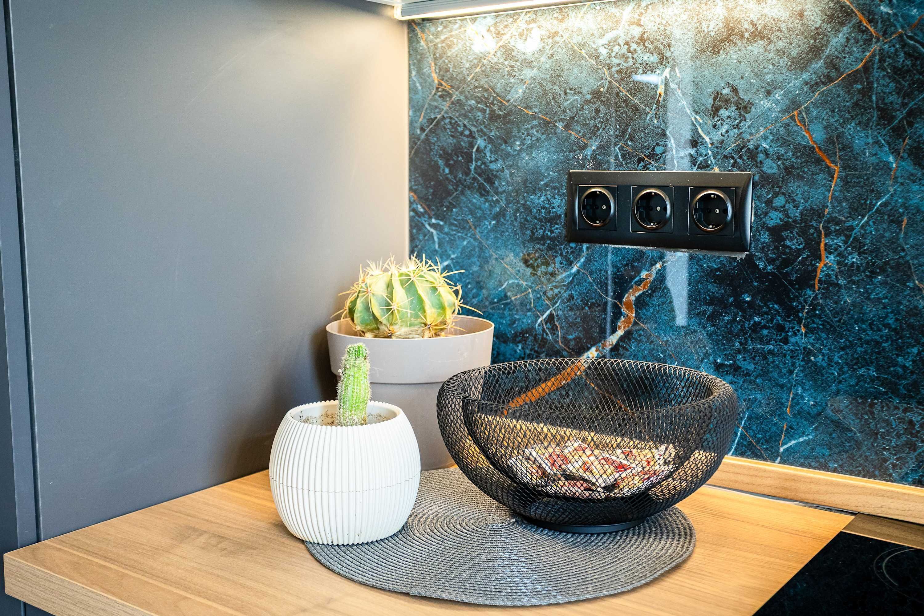 Een modern interieur met een houten tafel, een witte vaas met een cactuss, een zwart geweven kom, en een muur met een blauw marmerpatroon en een stopcontact.