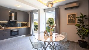 A modern kitchen with a built-in oven, a dining area with a round table and metal chairs, and a large window offering a view of greenery and a mountain. - Socrates Suites Open City Views in Kalamata (Kalamata)