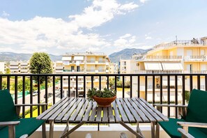 Ein Balkon mit Holztisch und -stühlen, einem Blumentopf und Blick auf Wohnhäuser und Berge.