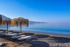 Ein Strand mit Liegestühlen und Strohsonnenschirmen, ein Holzdeck und ein ruhiges Meer unter blauem Himmel.
