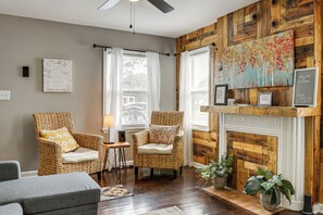 A room with two wicker chairs, a grey sofa, a fireplace with a mantel, and a wooden wall. - 4 Mi to Downtown Columbus! Updated Home w/ Yard (Columbus)