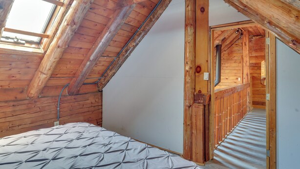 A wooden interior with a sloped ceiling, a bed with a patterned mattress, and a hallway leading to another room. - Classic New England Cabin on Belleau Lake (Wakefield)