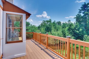 A wooden deck with a railing, a window with a fan, and a view of a forest. - Classic New England Cabin on Belleau Lake (Wakefield)