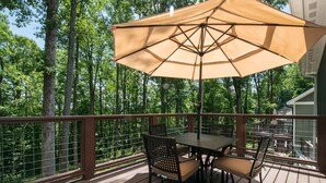 Terraza de madera con una mesa, sillas y un gran paraguas, con vista a un área boscosa.