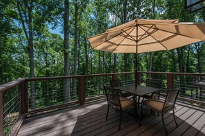 Um deck de madeira com mesa e cadeiras, ladeado por guarda-corpo de metal, com vista para uma floresta.