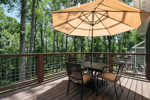A wooden deck with a table and chairs, a large umbrella, and a view of a forested area. - Druid Hills Retreat II Sleeps 8 Free Tix (Hendersonville)