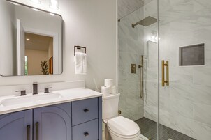 Un baño moderno con lavamanos, tocador y retrete blancos. Cuenta con una cabina de ducha de vidrio y un cabezal de ducha dorado.