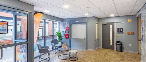 A modern interior with a herringbone wood floor, a seating area with a table and chairs, a potted plant, and a door with a fire alarm panel.
