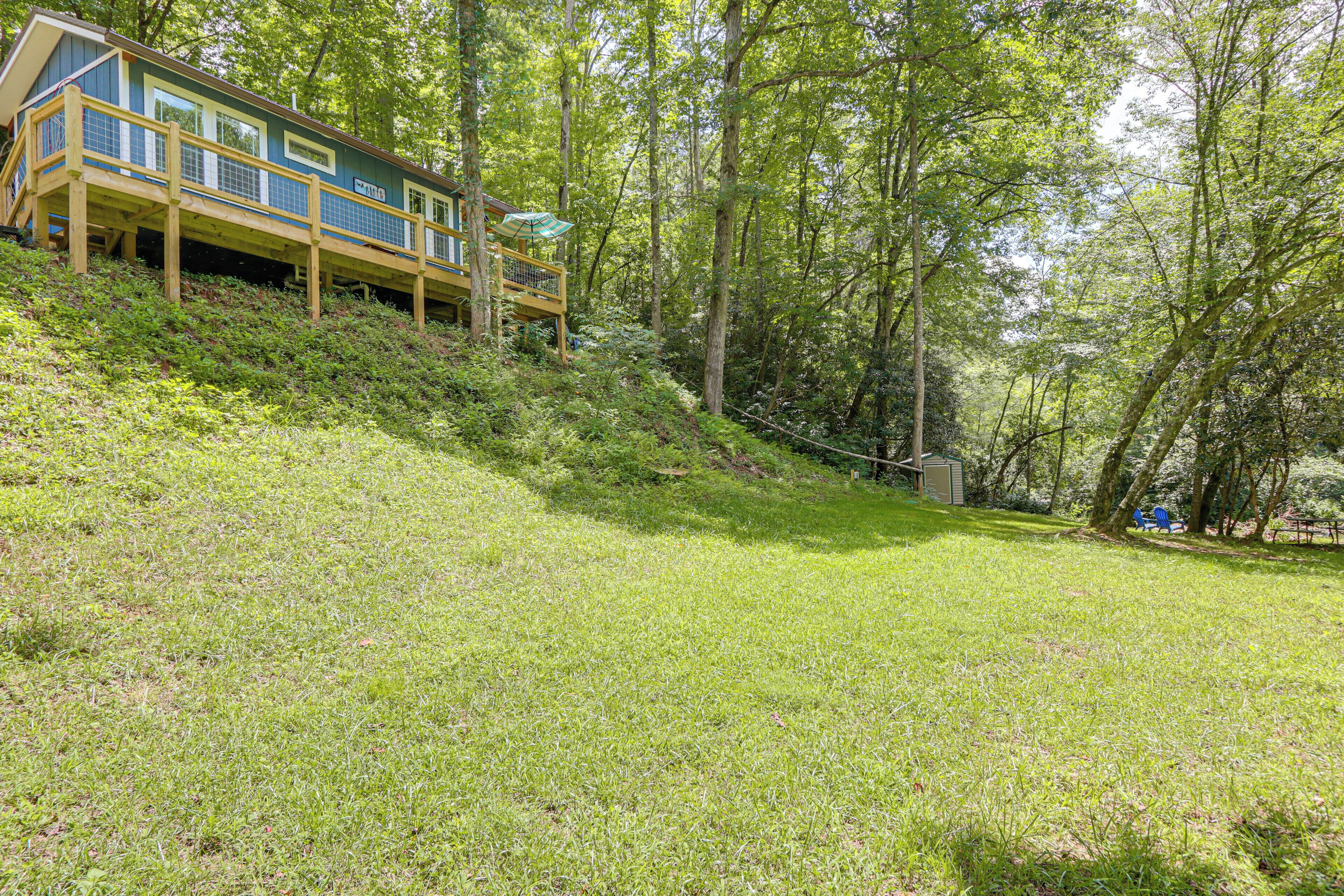 A house with a wooden deck and a covered area, surrounded by a grassy slope and trees.