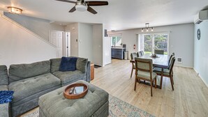 Un salon comprenant un canapé d’angle gris, une table basse avec un plateau, un coin repas avec une table et des chaises en bois, et un ventilateur de plafond.