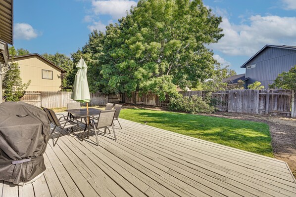 A backyard with a wooden deck, a barbecue grill, a table with chairs, and a large tree.