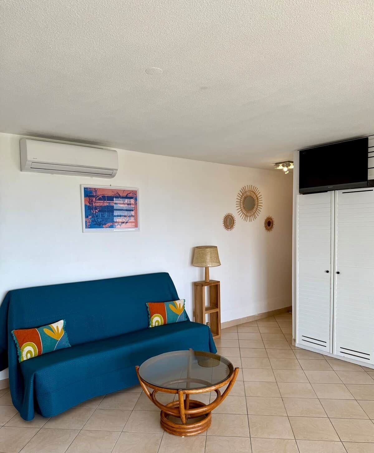 Una sala de estar con un sofá azul, una mesa de centro de vidrio, un televisor montado en la pared y un armario.