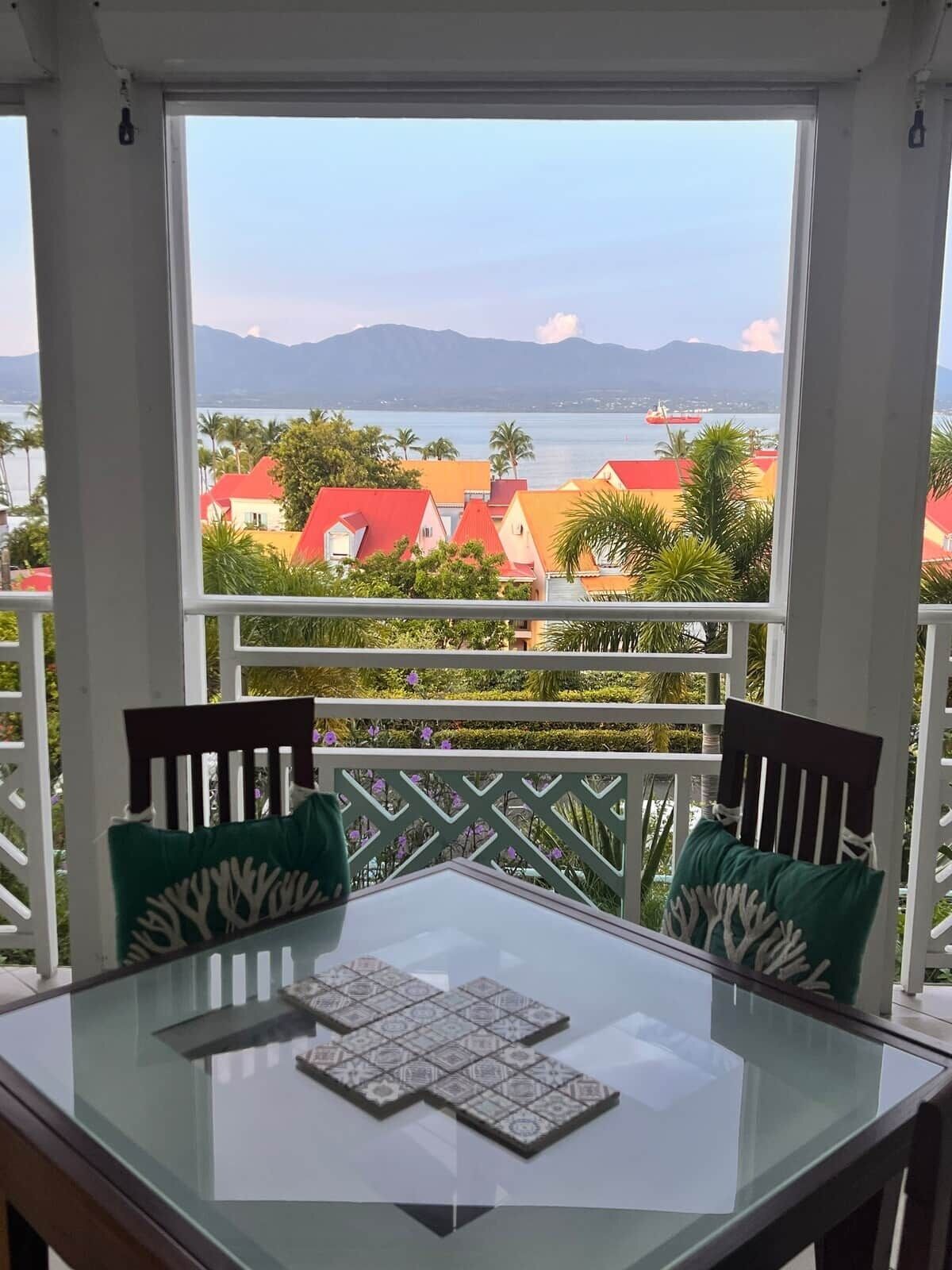 A balcony with a glass table and chairs, overlooking a coastal area with red-roofed buildings and a clear view of the sea and mountains.