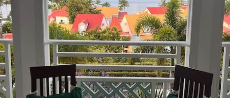 A balcony with a glass table and chairs, overlooking a coastal area with red-roofed buildings and a clear view of the sea and mountains.
