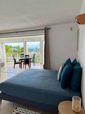 Ein Schlafzimmer mit Bett, Kissen, einem Nachttisch, einer Lampe und Blick auf einen Balkon mit Tisch und Stühlen.