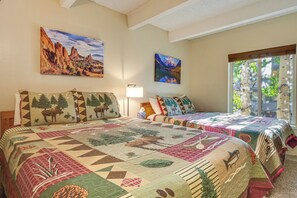 A room with two beds, each with a distinct quilt design, a window with a view of greenery, and two wall-mounted paintings. - Steps to the Slopes! Breck Ski Condo w/ Fireplace (Breckenridge)