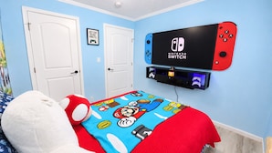 Una camera da letto con una Nintendo Switch montata a parete, un letto con coperta a tema Mario e un peluche.